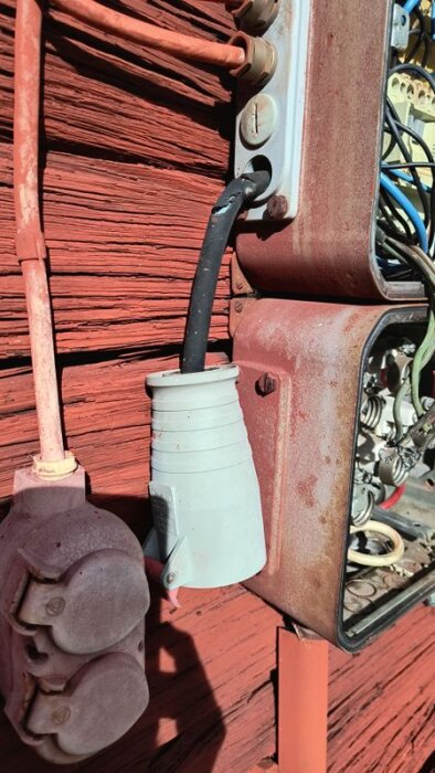 Röd byggnadsvägg med exponerad elektrisk installation, inklusive uttag och kabel. En del av kablaget är i dåligt skick och solbelyst.