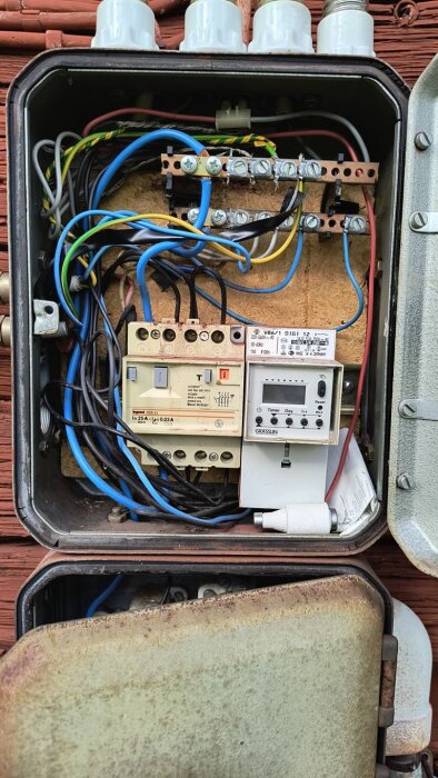 Elskåp med kablar och kopplingar, inklusive ett timerur och en jordfelsbrytare, innan byte av JFB, monterat på en röd vägg.