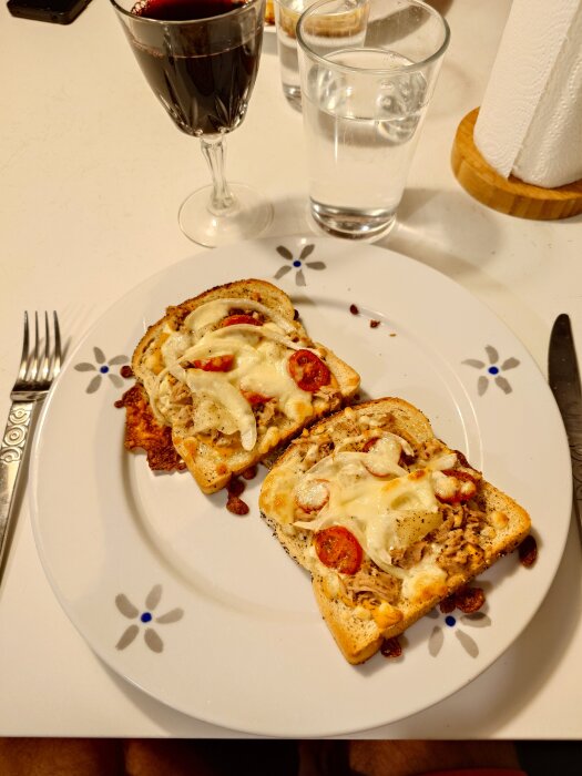 Varma mackor med majonäs, gul senap, tonfisk, lök, ananas, tomat och ost på tallrik, med glas rödvin och glas vatten i bakgrunden.