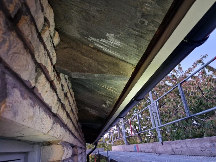 Närbild av takfotsbrädor och tegelvägg på en villa under renovering, med montering av hängränna och ställning i bakgrunden.