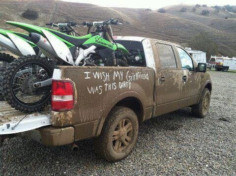 Lerig pickup med motorcyklar bakpå, text på sidan lyder: "I wish my girlfriend was this dirty." Parkerad på grusväg med kullar i bakgrunden.