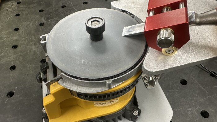 Slipmaskin med roterande skiva för verktygsslipning, med röd bryningsjigg ovanpå aluminiumextrabordet för att hålla verktyget plant.