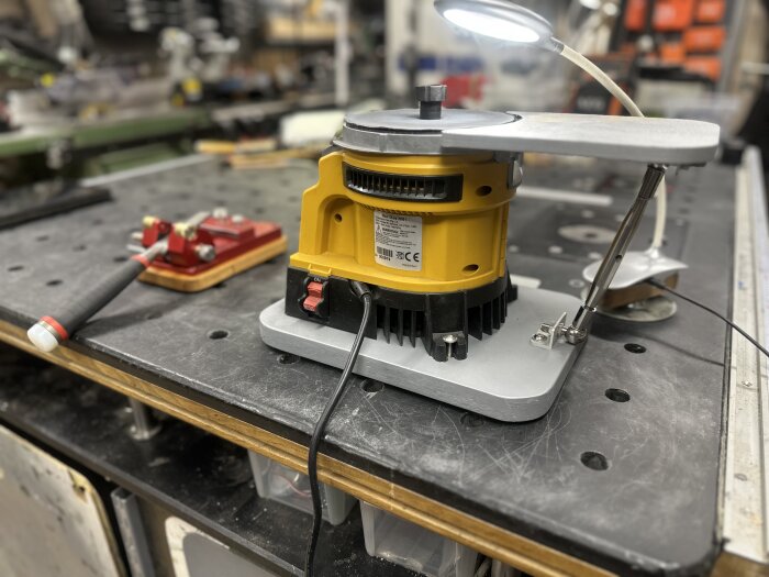 Gul slipmaskin med extrabord placerad på en arbetsbänk; justerbar lampa och röd jiggsupport synlig bredvid.