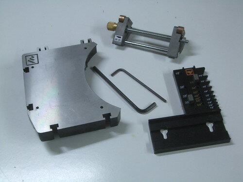 Delar till ett justerbart extrabord för slipmaskin, inklusive aluminiumskiva, vinkelstöd och monteringsverktyg, ligger på ett vitt bord.