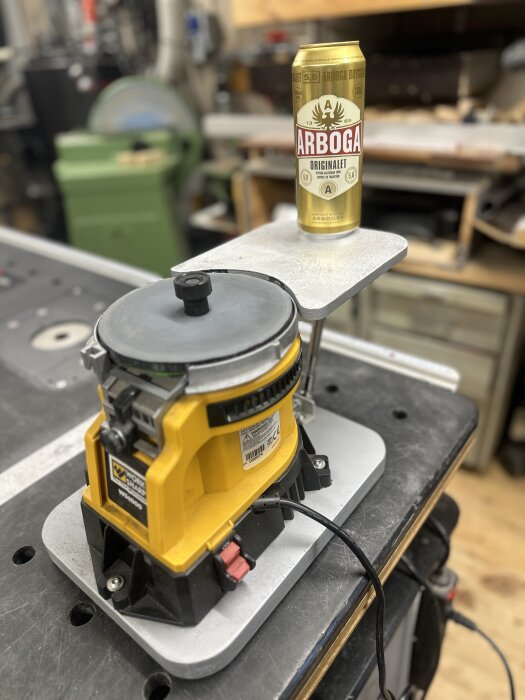 En slipmaskin med gult hölje, monterad på ett arbetsbord med ett extrabord, en ölburk med texten "Arboga" placerad ovanpå.