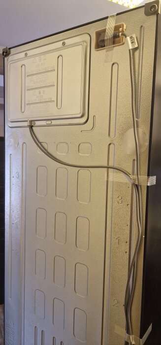 Baksidan av ett kylskåp med kabelanslutningar och varningstexter, monterat med några tejpbitar.