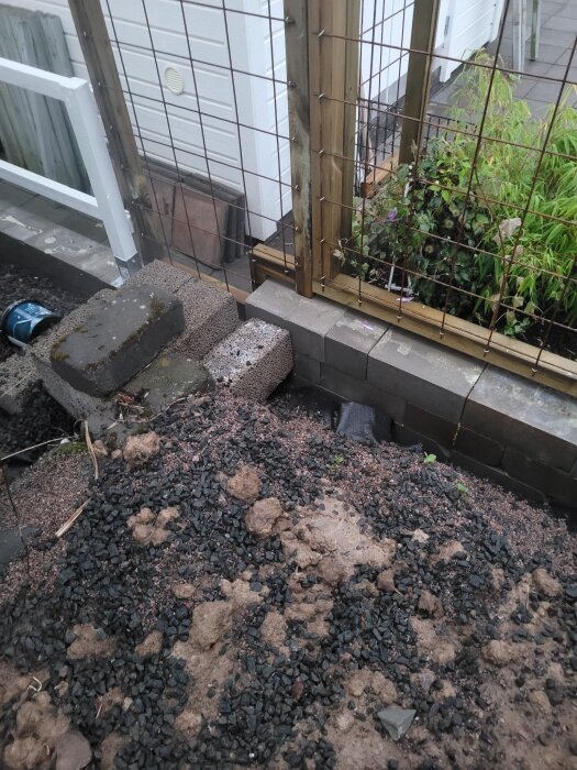 Byggmaterial vid stängsel mot hus, staplade block och jord. En del av stängslet saknas för solplats bakom. Grön växt intill träram.
