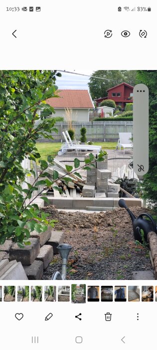 Trädgård med byggmaterial, staplade plattor och en vattenkanna. I bakgrunden två solstolar vid ett trädäck, gränsat av staket och träd.