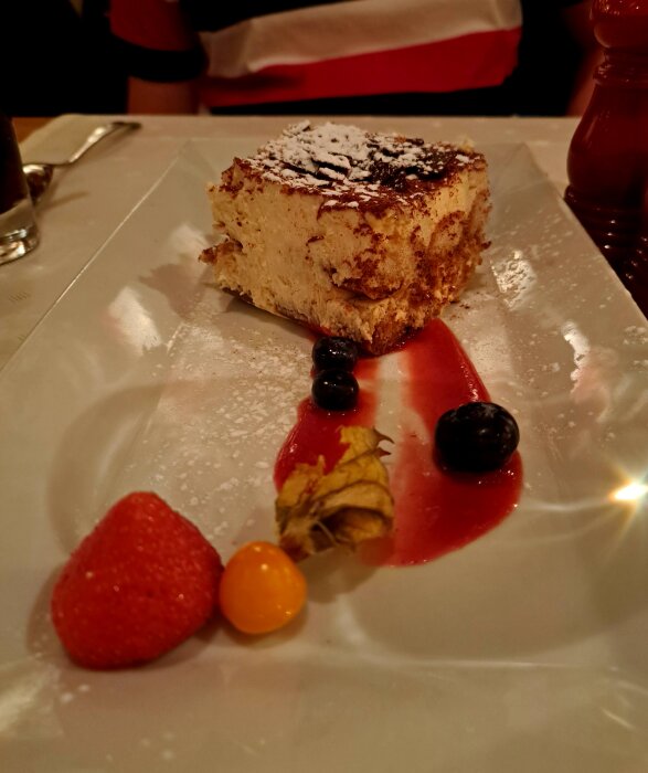 Tiramisu på rektangulärt fat med jordgubbe, physalis och blåbär, garnerat med florsocker och röd sås.