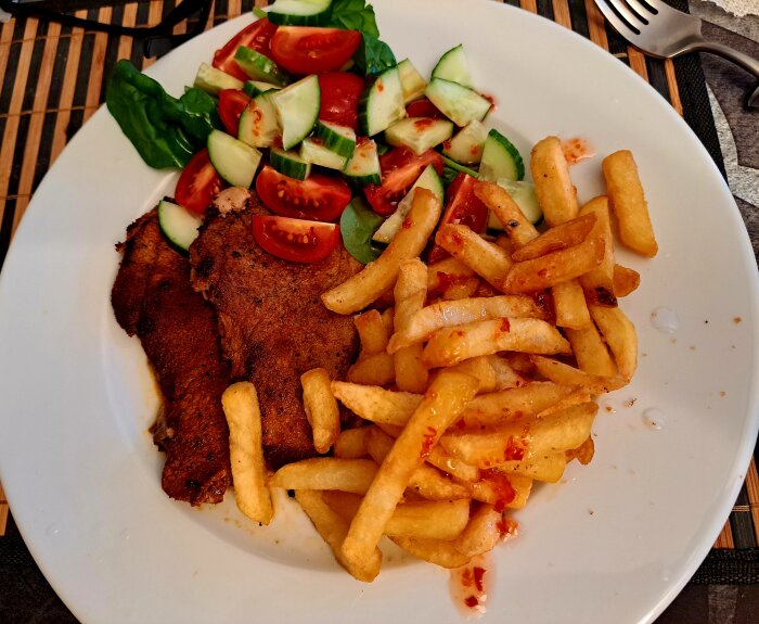 En tallrik med kött, krispiga pommes frites med chilisås, samt en fräsch sallad med gurka, tomat och bladgrönsaker.