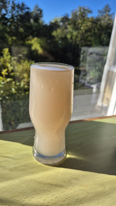 Ett glas med grumligt vatten och mycket sediment står på ett bord vid ett fönster med träd i bakgrunden.