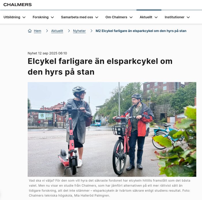 Två personer med hjälmar står på en asfalterad väg, en med elsparkcykel och en med elcykel, utanför byggnader och gröna träd.
