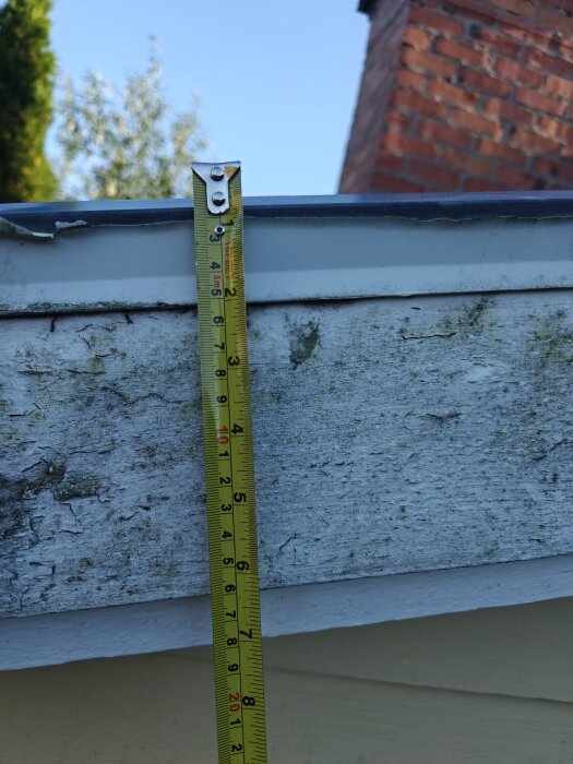 Måttband mot fasadplanka på takkupa, mäter höjd upp till drygt 17 cm, med tegelvägg i bakgrunden.