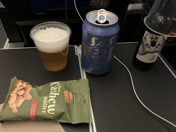 Plastglas med öl, burk med SAS IPA, flaska vin med koalaetikett och påse cashewnötter på ett flygplansbord.