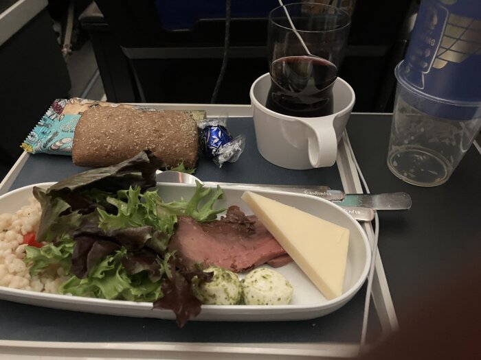 Flygplansmåltid med sallad, rostbiff, ostskiva, pärlcouscous och bröd, serverad med ett glas rött vin och en kaffekopp på ett brickbord.