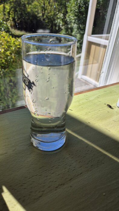 Glas med klart vatten på ett bord, solbelyst bakgrund av trädgård.