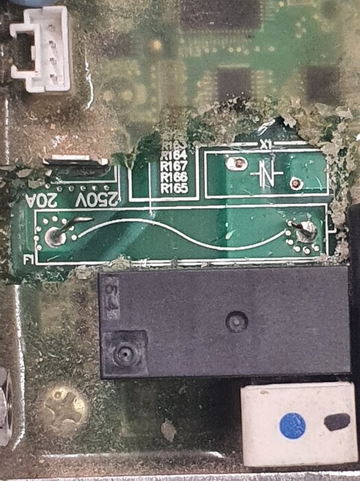 Kretskort med texten "250V 20A" och "X1", närbild på komponenter och lödningar, delvis skadad eller smutsig yta.