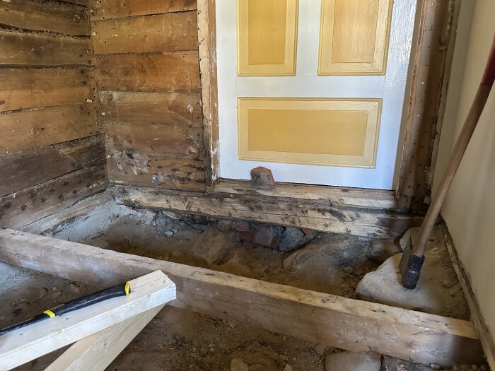 Renovering av äldre kök, synligt bjälklag, trädörr med gul panel, verktyg på golvet, jord och stenexponering.