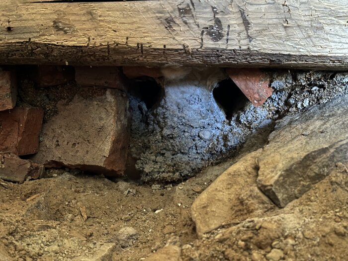 Hål i grund med tegelstenar och bjälke, delvis fylld med sand och jord, visar plats för avloppsinstallation och rördragning.