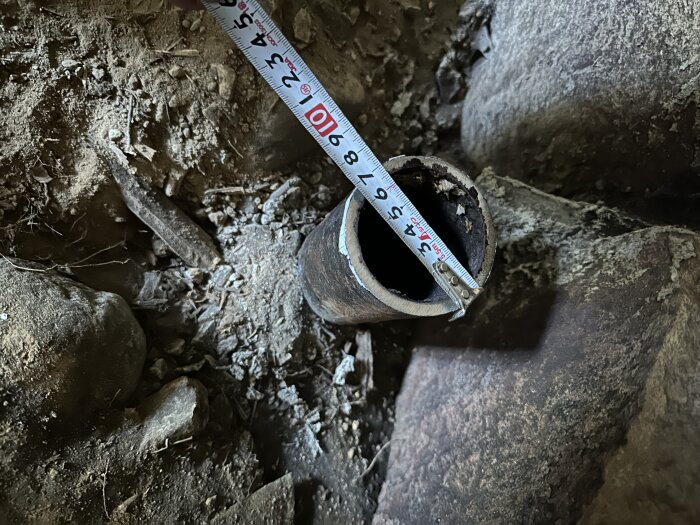 Gjutjärnsrör i marken med ett måttband som visar 70 mm ytterdiameter; omgivande jord och stenar syns.