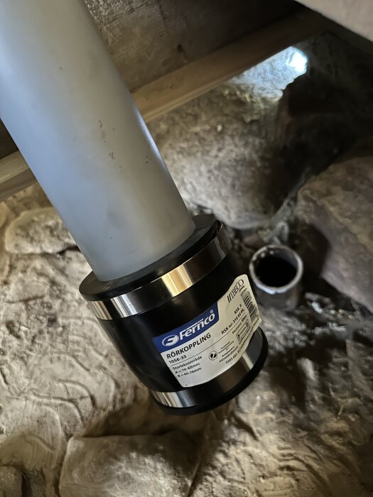 Fernco rödkoppling installerad på ett vitt plaströr, placerad i ett kryputrymme med murad stenvägg i bakgrunden.