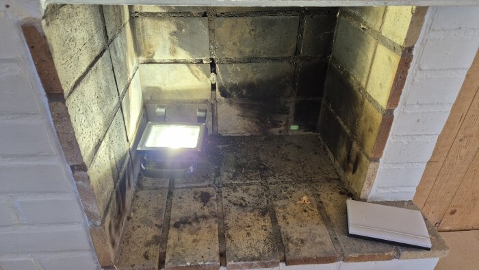 Gammal eldstad med smutsig insida, tegelväggar och en ljuskälla som lyser upp insidan av spisen.