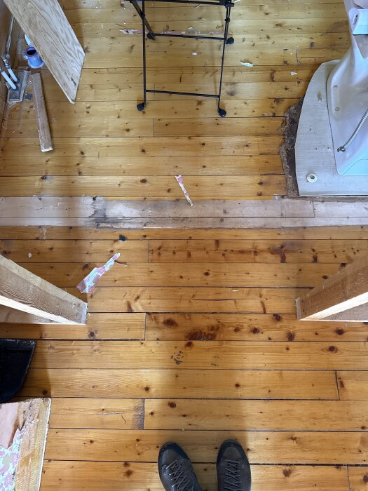 Renovering av ett gammalt trägolv i badrum, med slitna plankor och byggmaterial runtomkring. En person står i bildens nedre del.