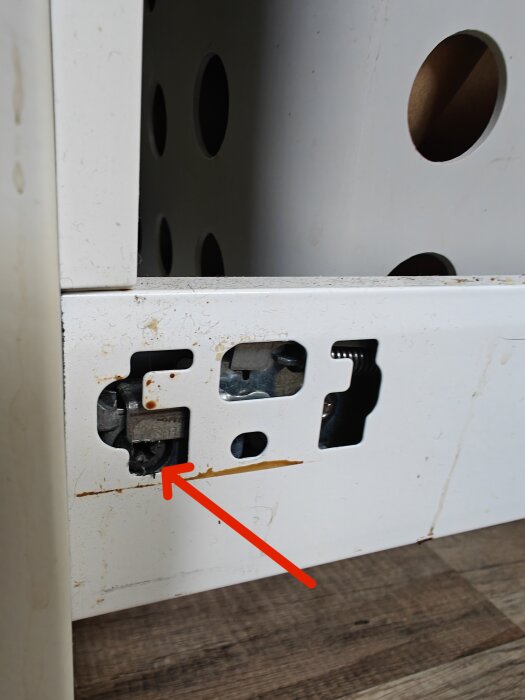 Närbild på en justeringsmekanism med rostfläckar på möbelbeslag, markerad med en röd pil, synlig inuti en vit IKEA-möbel.