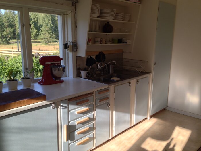 Kök med 50-talsinredning, blå underskåp och vita övre hyllor. Röd köksassistent på bänkskivan bredvid diskho. Fönster mot trädgård i bakgrunden.