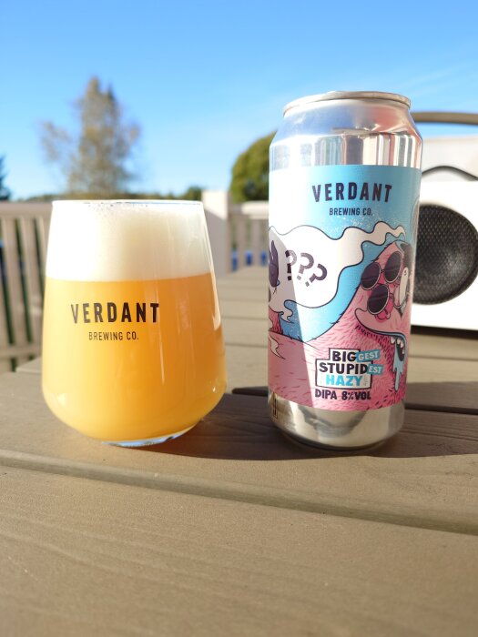 Glas med Verdant öl bredvid en Verdant burk, med blå himmel och utemöbler i bakgrunden.