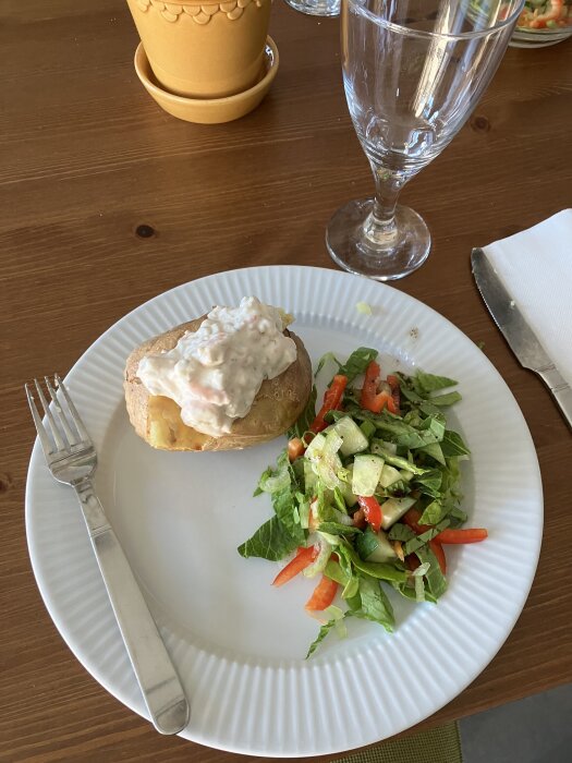 Bakad potatis med skagenröra och extra räkor samt en grönsallad på en tallrik med gaffel och glas.