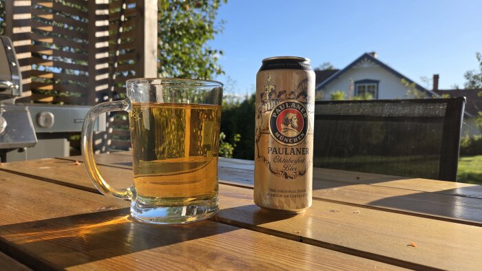 En ölsejdel och en burk Paulaner Oktoberfest Bier på ett utebord, solbelyst på sensommaren med en grill och ett hus i bakgrunden.