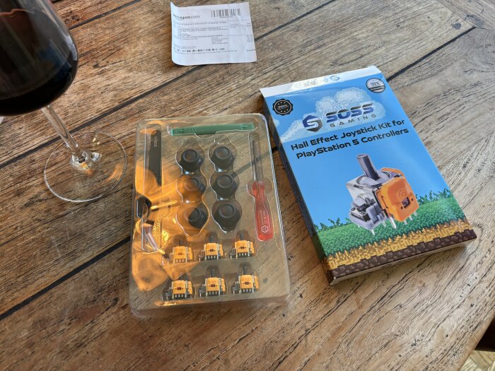 PlayStation 5 joystick kit och verktyg på träbord, bredvid ett glas vin och en Amazon-leverans.