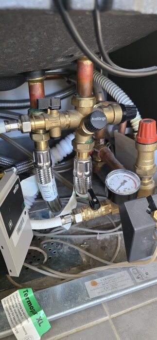 Värmesystem med ventil och tryckmätare under en värmepump, omgiven av kopparrör och flexibla slangar.
