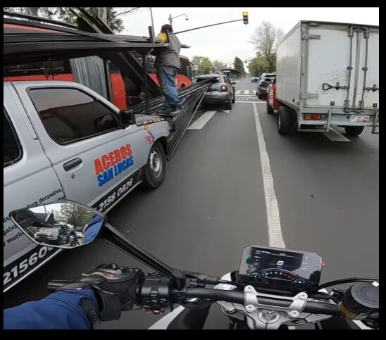 Motorcyklist kör på väg bakom pickup lastad med metallrör och en vit lastbil, med trafikljus och flera fordon framåt.