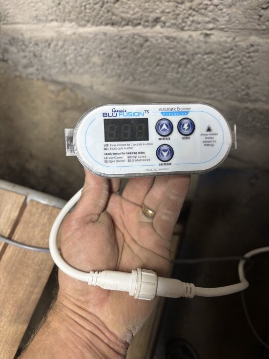 En hand håller en Balboa 5-polig enhet med texten "Genesis Blu Fusion TS Automatic Bromine Generator" och en vit kabel och kontakt.