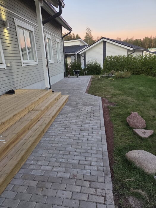Hus med grå fasad, trätrappa och stenlagd gång. Gräs och stenar syns vid sidan om. Solnedgång i bakgrunden. Trädgårdsarbete pågår.