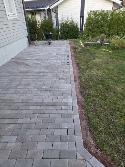 Stenlagd uteplats med marksten bredvid en gräsmatta, med en skottkärra i bakgrunden och ett hus synligt.