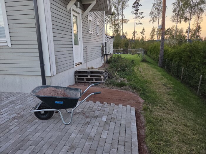 Byggarbetsplats vid hus med skottkärra fylld med sand på stenlagd gång och murpallar vid dörren, omgiven av gräs och träd.