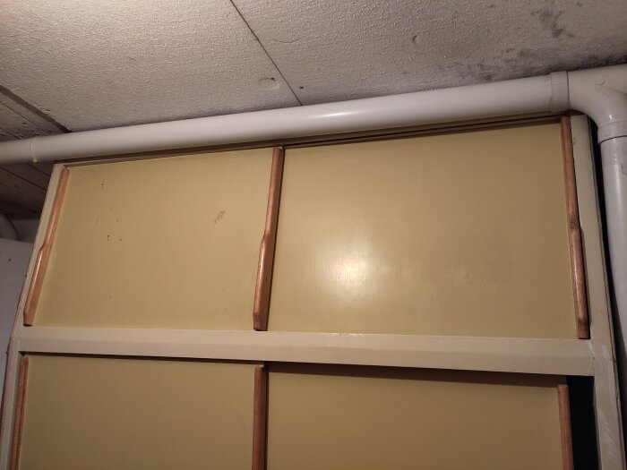 Gul dörr med trälister och handtag på båda sidor, rör vid taket, i ett källarutrymme.