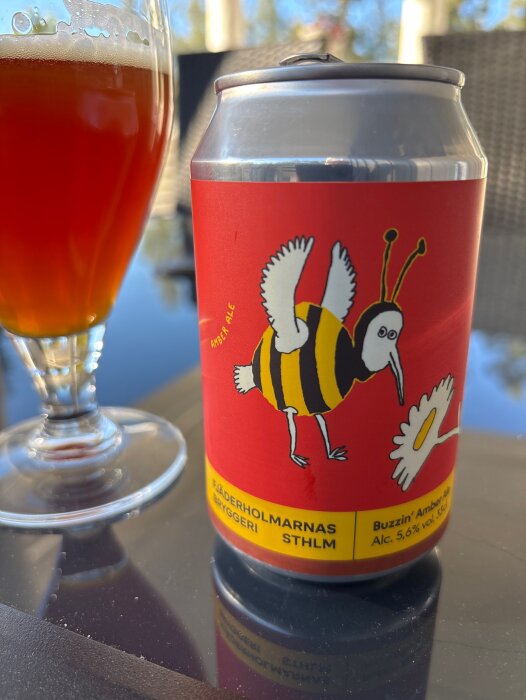 Burk med Fjäderholmarnas Buzzin' Amber Ale och ett glas öl på ett bord i solen.