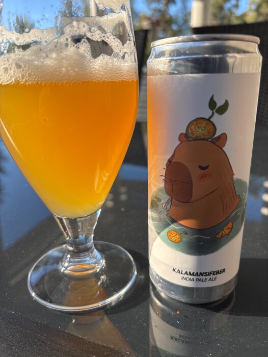 Glas med bärnstensfärgad öl och burk med etikett föreställande en tecknad figur och texten "Kalamansifeber India Pale Ale" på ett bord i solen.