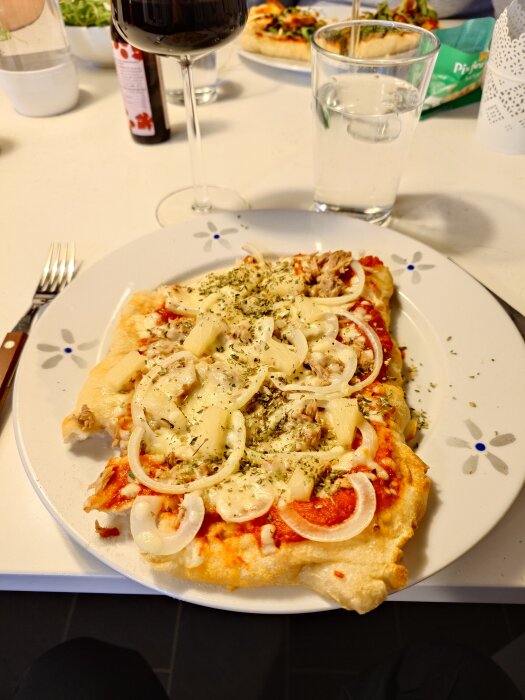 Pinsa toppad med tonfisk, lök och ananas på en tallrik i närbild, ett glas rött vin och vatten syns i bakgrunden.