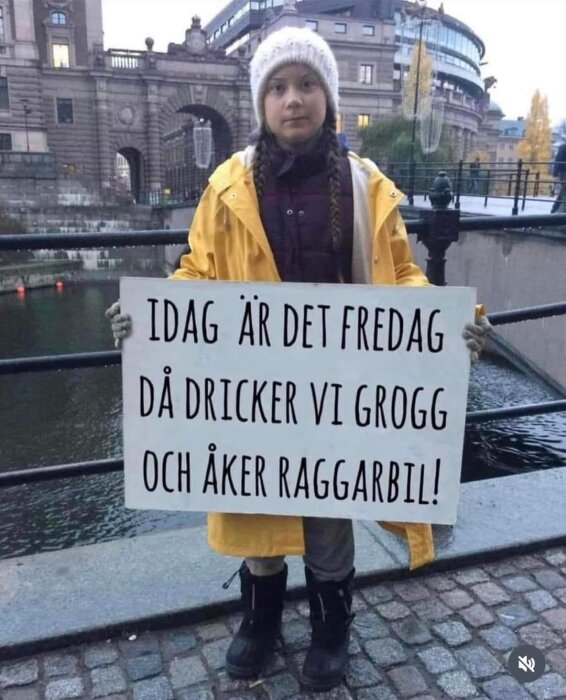 Person i gul regnjacka och vit mössa håller en skylt vid en kanal. Text på skylten: "IDAG ÄR DET FREDAG DÅ DRICKER VI GROGG OCH ÅKER RAGGARBIL!