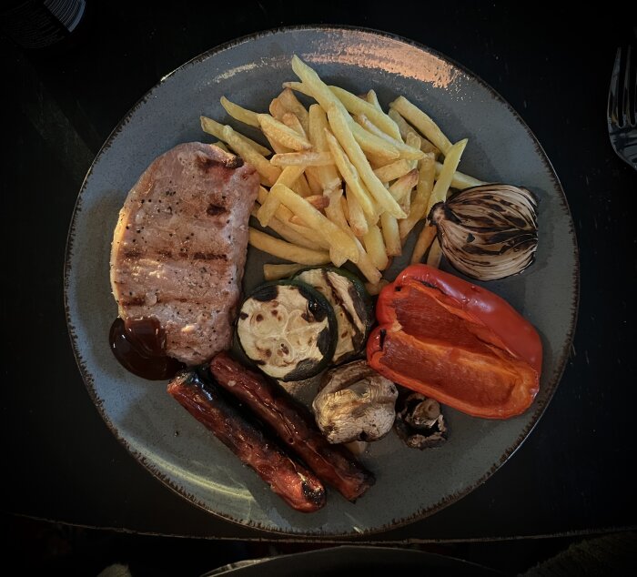 Grillad fläskkotlett, korv, paprika, champinjoner, lök, squash med pommes och BBQ-sås på tallrik.