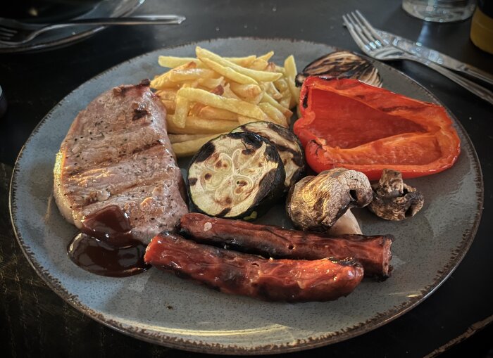 Grilltallrik med korv, fläskkotlett, röd paprika, champinjoner, lök, squash, pommes frites och BBQ-sås på en tallrik.