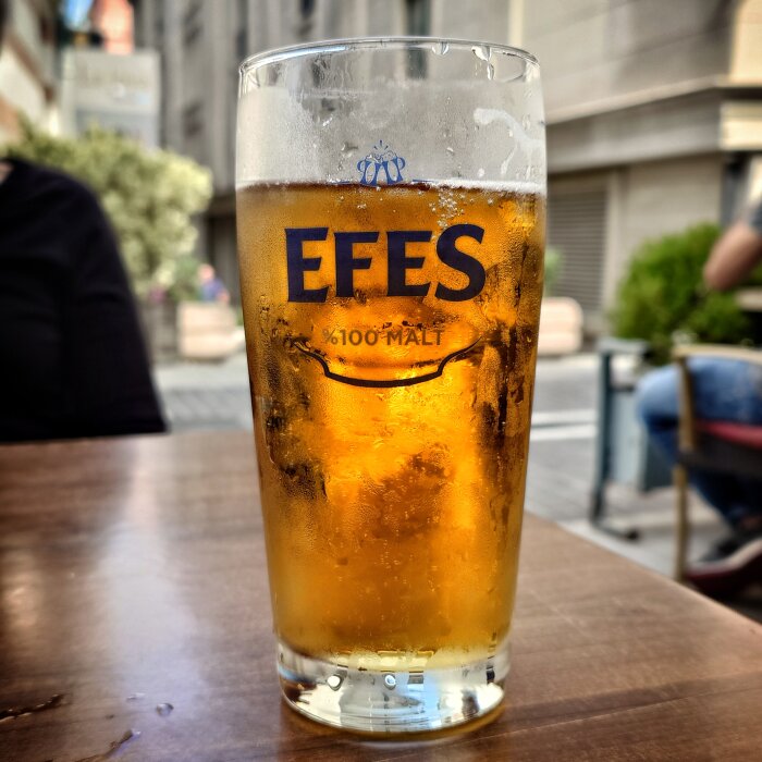 Ett glas öl med texten "EFES" på glaset, placerat på ett träbord utomhus med en suddig bakgrund av en gata.