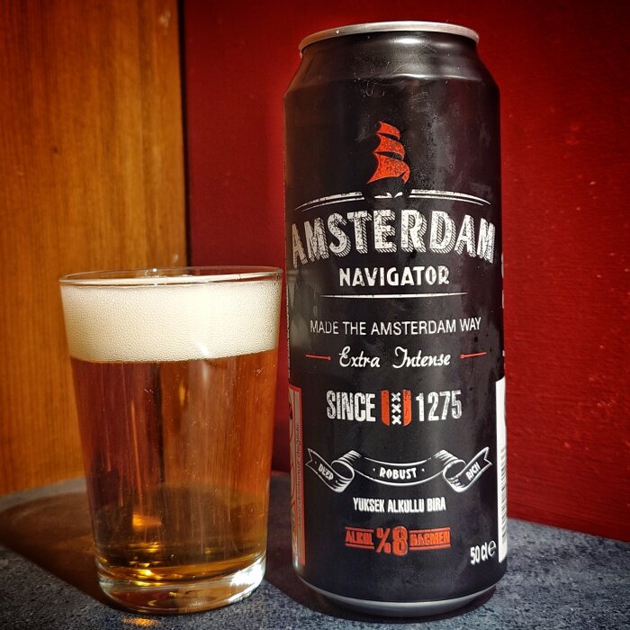 En burk Amsterdam Navigator öl står bredvid ett glas med öl mot en röd och träbakgrund.