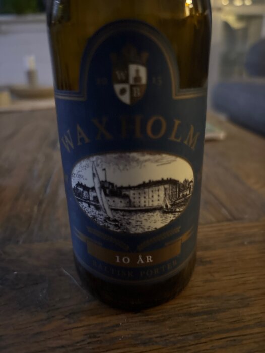 Flaska med etikett "Waxholm 10 år" och motiv av båtar och byggnader, står på ett träbord.