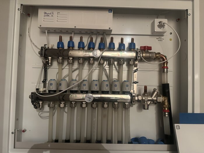 Golvvärmefördelare med flera blå ventiler och rör anslutna i en vit ram.